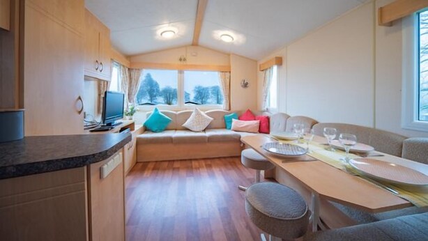 Interior compacto de una caravana con kitchenette, comedor y zona de estar con vistas a árboles.
