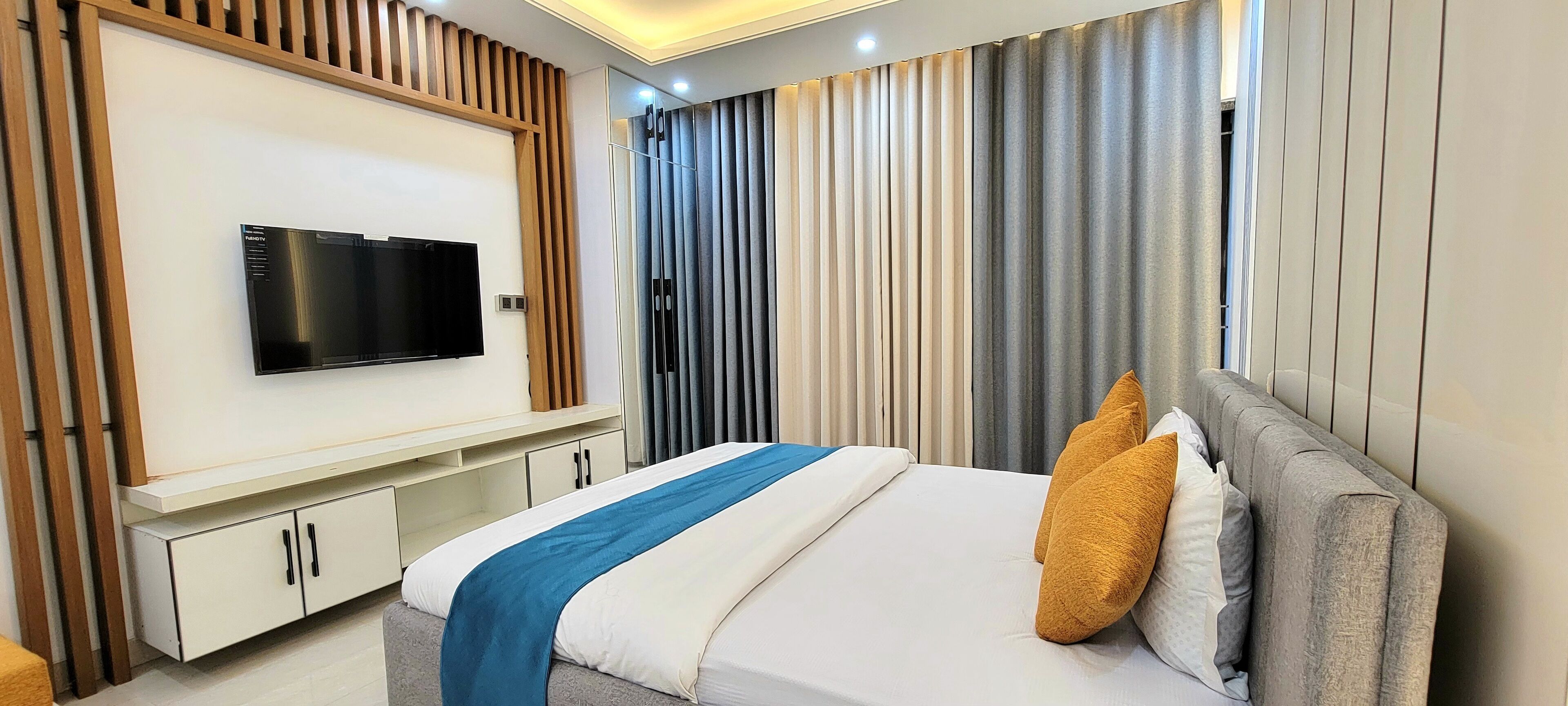 Een moderne hotelkamer met een flatscreen televisie bevestigd aan een houten paneel, een bed met een blauw-witte dekbedovertrek en een grijs hoofdeinde.