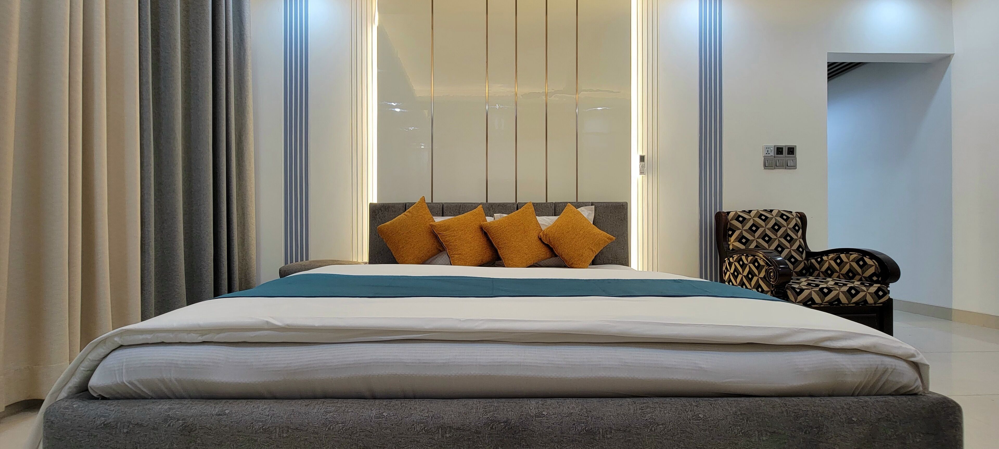 Un dormitorio moderno con una cama grande, un cabecero con muchos cojines, un sillón estampado y una puerta corrediza de cristal.