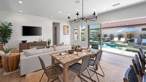 A modern living room with a dining area, a TV mounted on the wall, and a view of a pool and outdoor seating area. - Desert pool retreat w/ mountain views (La Quinta)
