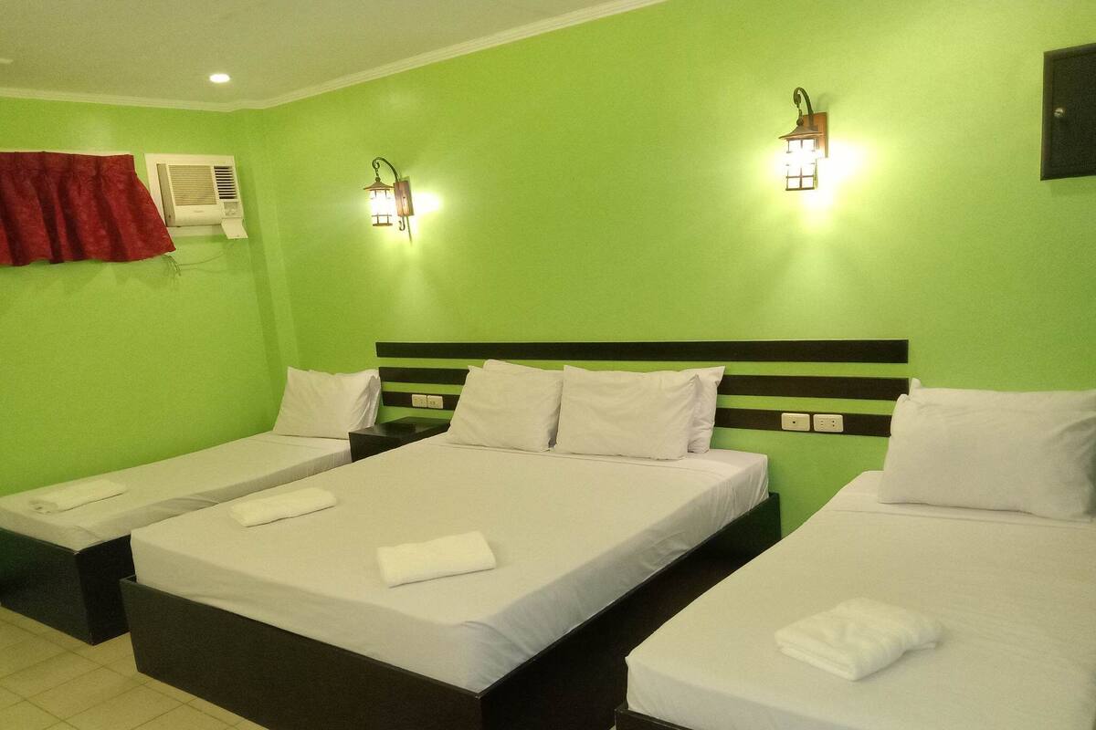 Habitación de hotel con tres camas individuales, cada una con ropa blanca y almohadas. Hay dos lámparas de pared a cada lado de una pared verde.
