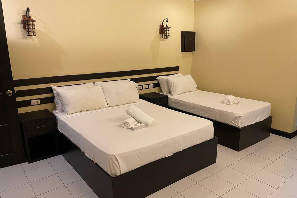 Una habitación de hotel con dos camas, cada una con ropa blanca y una toalla enrollada. Hay dos mesitas de noche, una a cada lado de las camas. La habitación tiene una pared color beige y un cabecero negro.