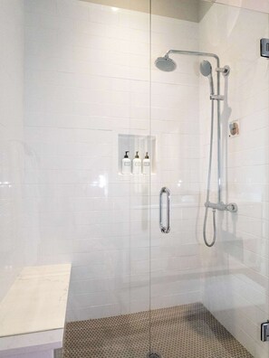 A glass-enclosed shower with a handheld showerhead, a wall-mounted faucet, and a control panel with two bottles.