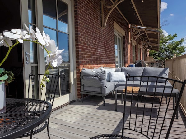 A patio with a metal table and chairs, a grey sofa, and a brick building with large windows.
