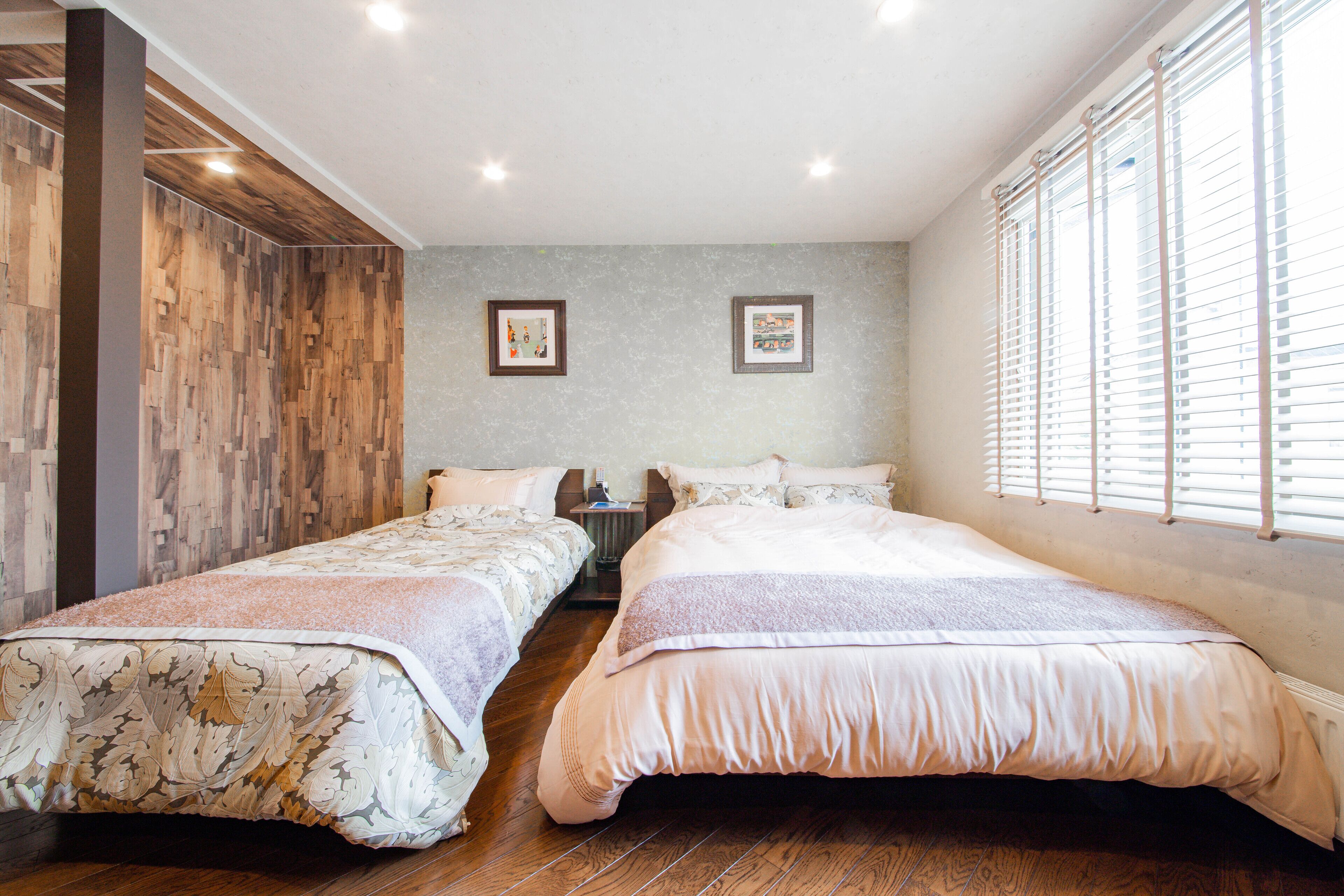A bedroom with two single beds, wooden flooring, a grey accent wall, and a window with blinds.