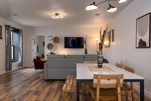 Una sala de estar moderna con un sofá gris, una mesa de comedor con sillas de madera y un televisor montado en la pared.