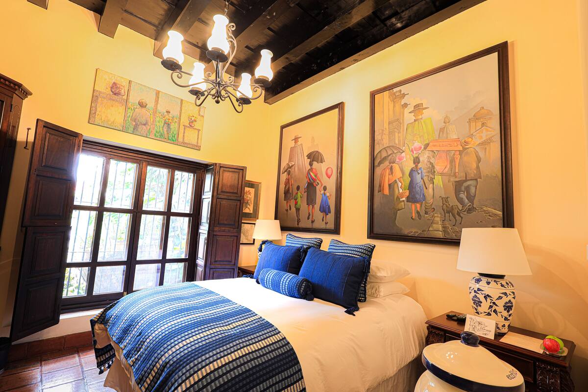 Una habitación de hotel con una cama grande, un candelabro, obras de arte en las paredes y una ventana con cortinas.