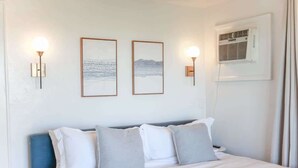 Un lit bien fait, avec une literie blanche et grise, deux œuvres d’art encadrées accrochées au mur et un climatiseur fixé au mur.