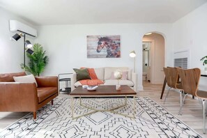 Uma sala de estar com sofá, mesa de centro e cadeiras, um quadro na parede e um aparelho de ar condicionado.