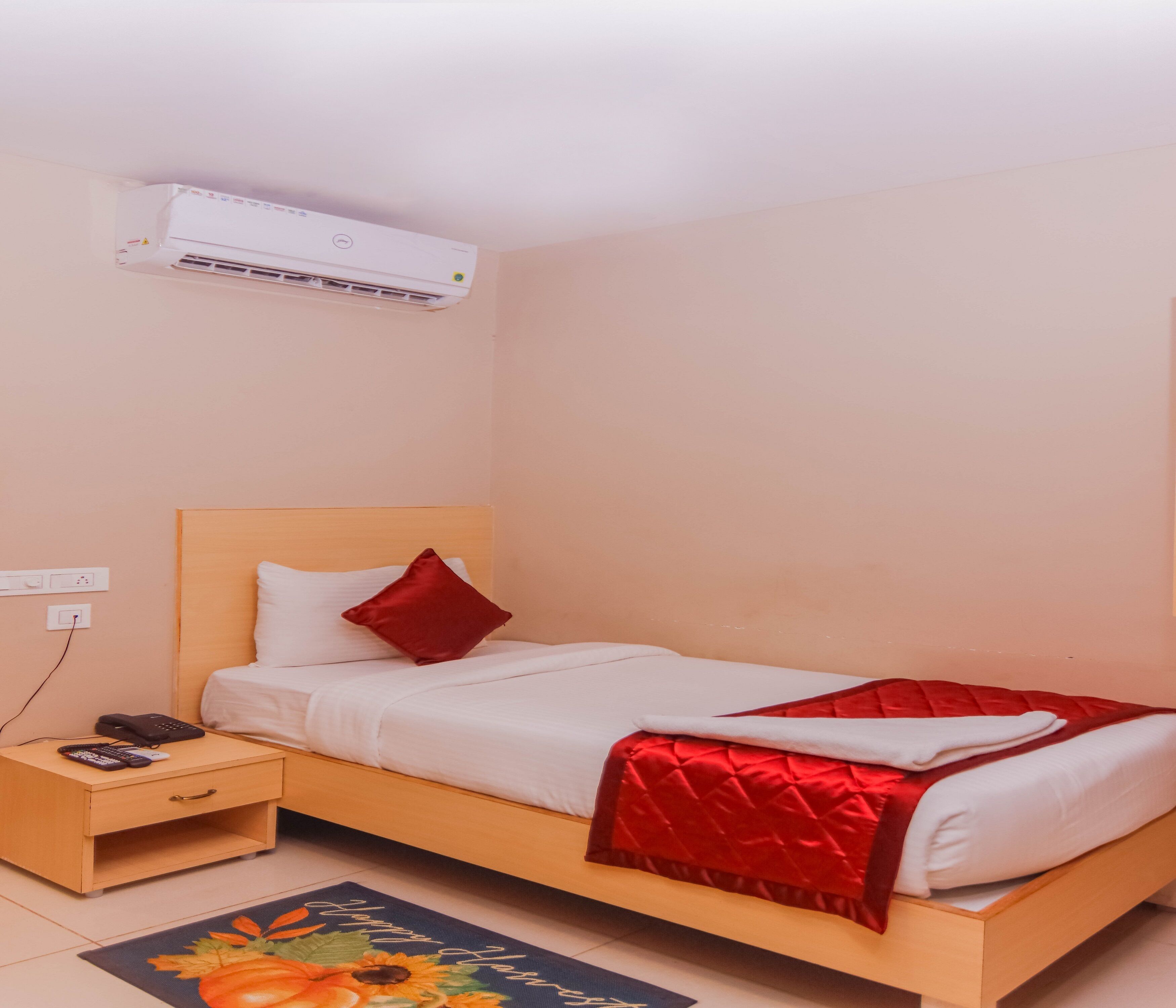 Un lit simple avec une tête de lit en bois, une couverture rouge et un oreiller blanc. Il y a une table de chevet avec un téléphone et une télécommande. Un appareil de climatisation est fixé au mur au-dessus du lit.