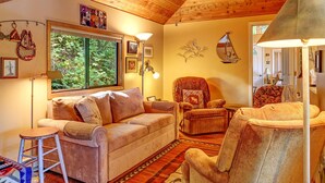 A cozy living room with a wooden ceiling, a large window, a sofa, a chair, and a lamp.