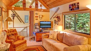 A cozy living room with a fireplace, a television, and a large window overlooking a forest.
