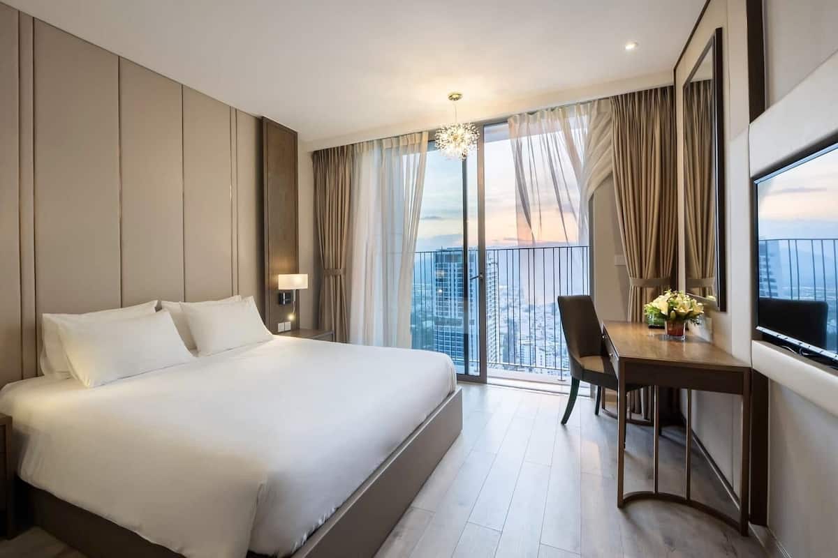A hotel room with a large bed, a desk with a chair, a TV, and a balcony with a city view.