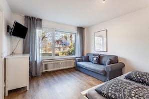 Quarto moderno com sofá cinza, cama com almofadas estampadas, televisão montada e janela com vista para uma casa.