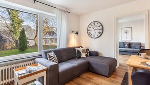 Un salon moderne avec un canapé d’angle gris, une table basse en bois, une horloge au mur et une vue sur l’extérieur.