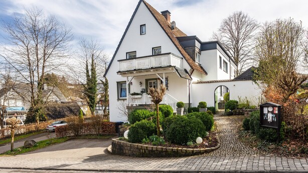Une maison à deux étages, dotée d’un toit en pente, d’un balcon et d’un jardin avec une allée pavée.