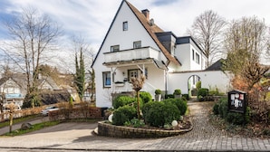 Ein zweigeschossiges Haus mit einem geneigten Dach, einem Balkon und einem Garten mit gepflasterter Auffahrt.