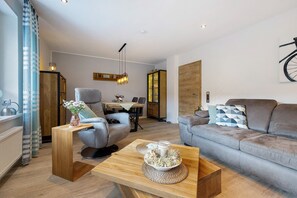 Una sala de estar moderna con sofá, sillón, mesa de centro de madera y un área de comedor al fondo.