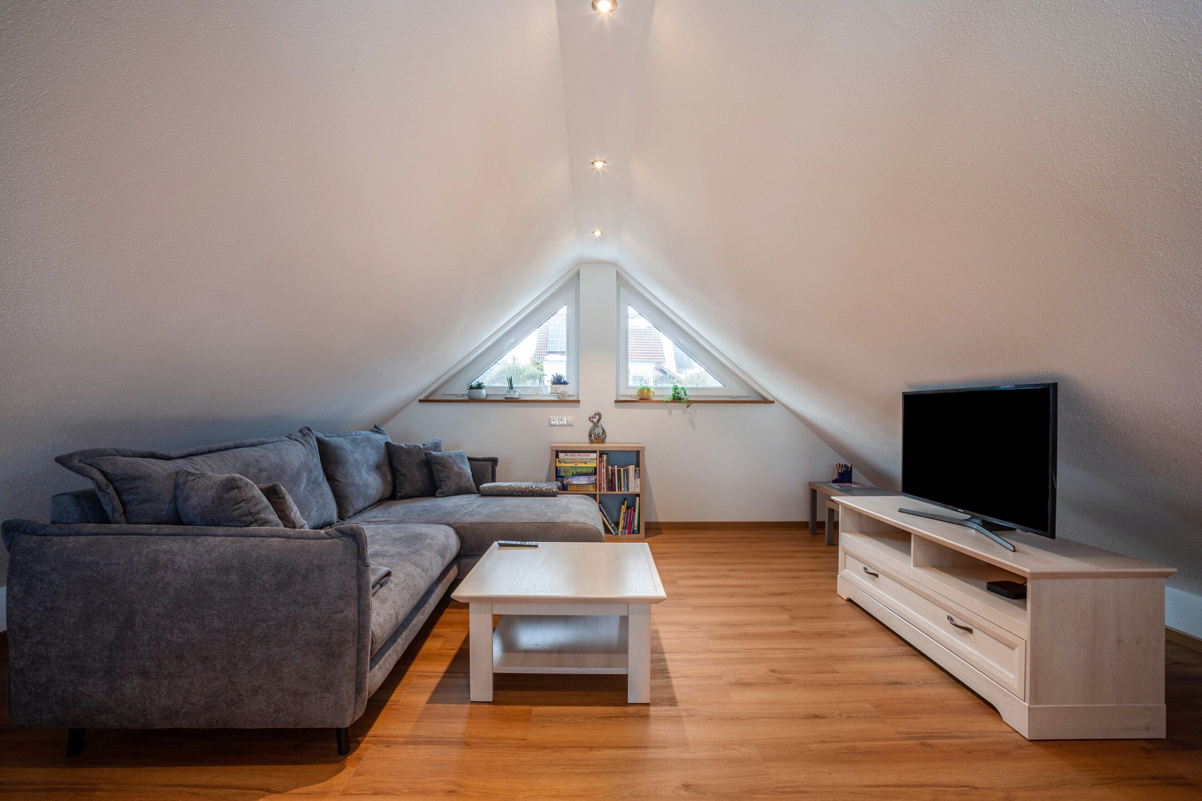 A modern loft apartment with a sloped ceiling, a grey sectional sofa, a white coffee table, a wooden TV stand, and a bookshelf.