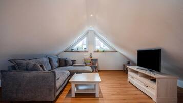 A modern loft apartment with a sloped ceiling, a grey sectional sofa, a white coffee table, a wooden TV stand, and a bookshelf.