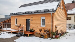A wooden cabin with a snow-covered roof, a wooden porch, and potted plants.
