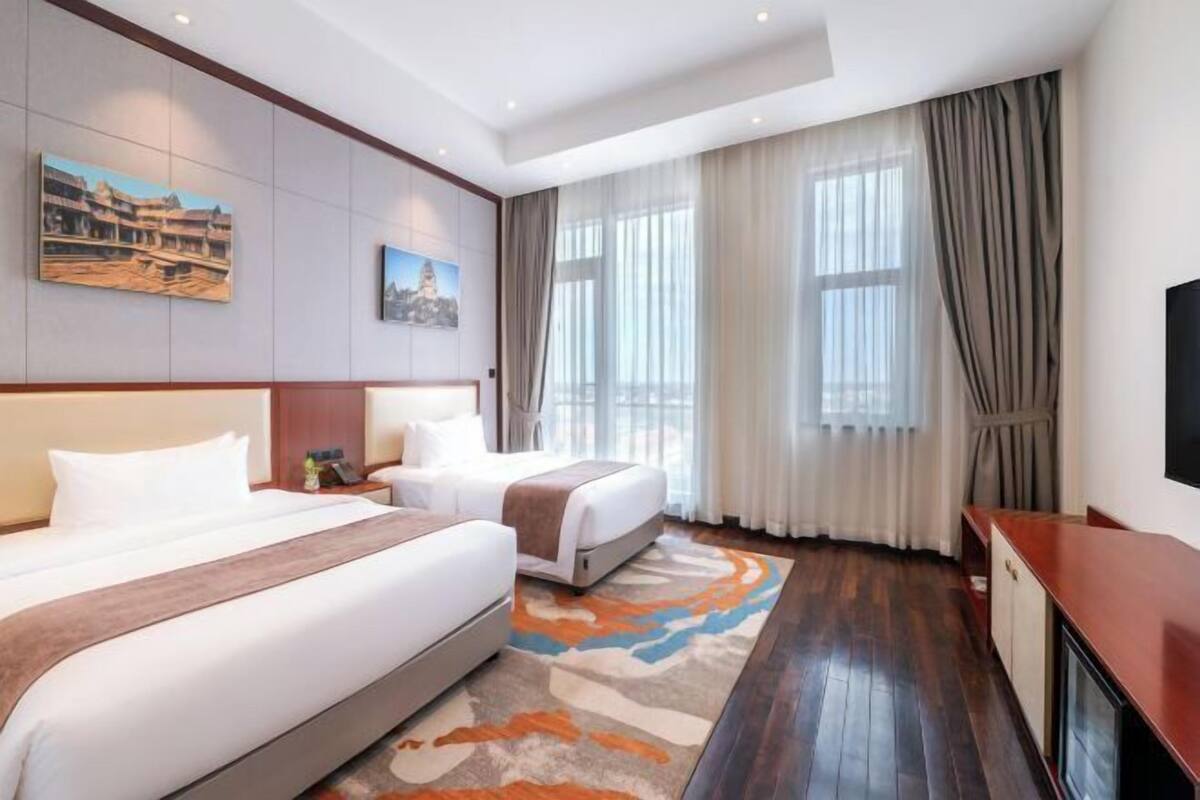 Een hotelkamer met twee bedden, een groot raam met gordijnen, een flatscreen-televisie en kunstwerken aan de muren.