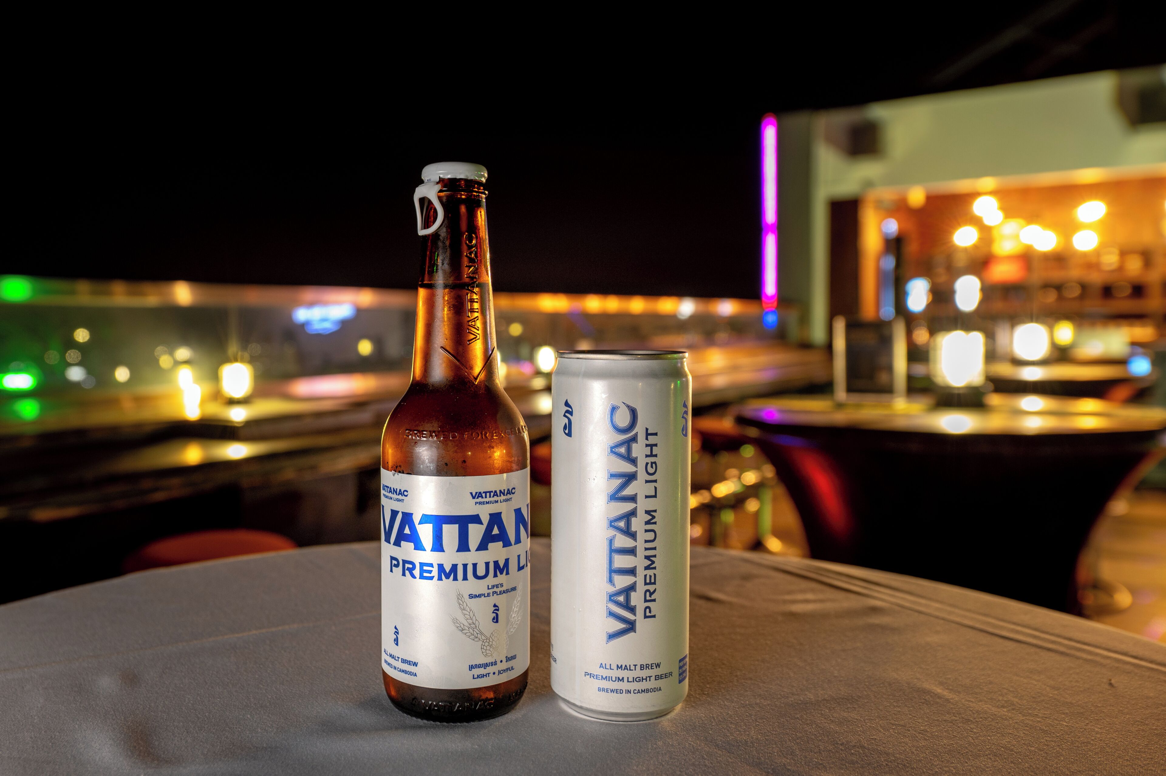 A bottle and a can of Vattanac Premium Light beer on a table with a cityscape view in the background.