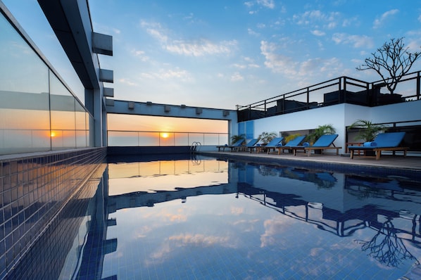 Une piscine à l’eau d’un bleu cristallin, le soleil couchant en toile de fond et des chaises longues disposées le long du bord de la piscine.