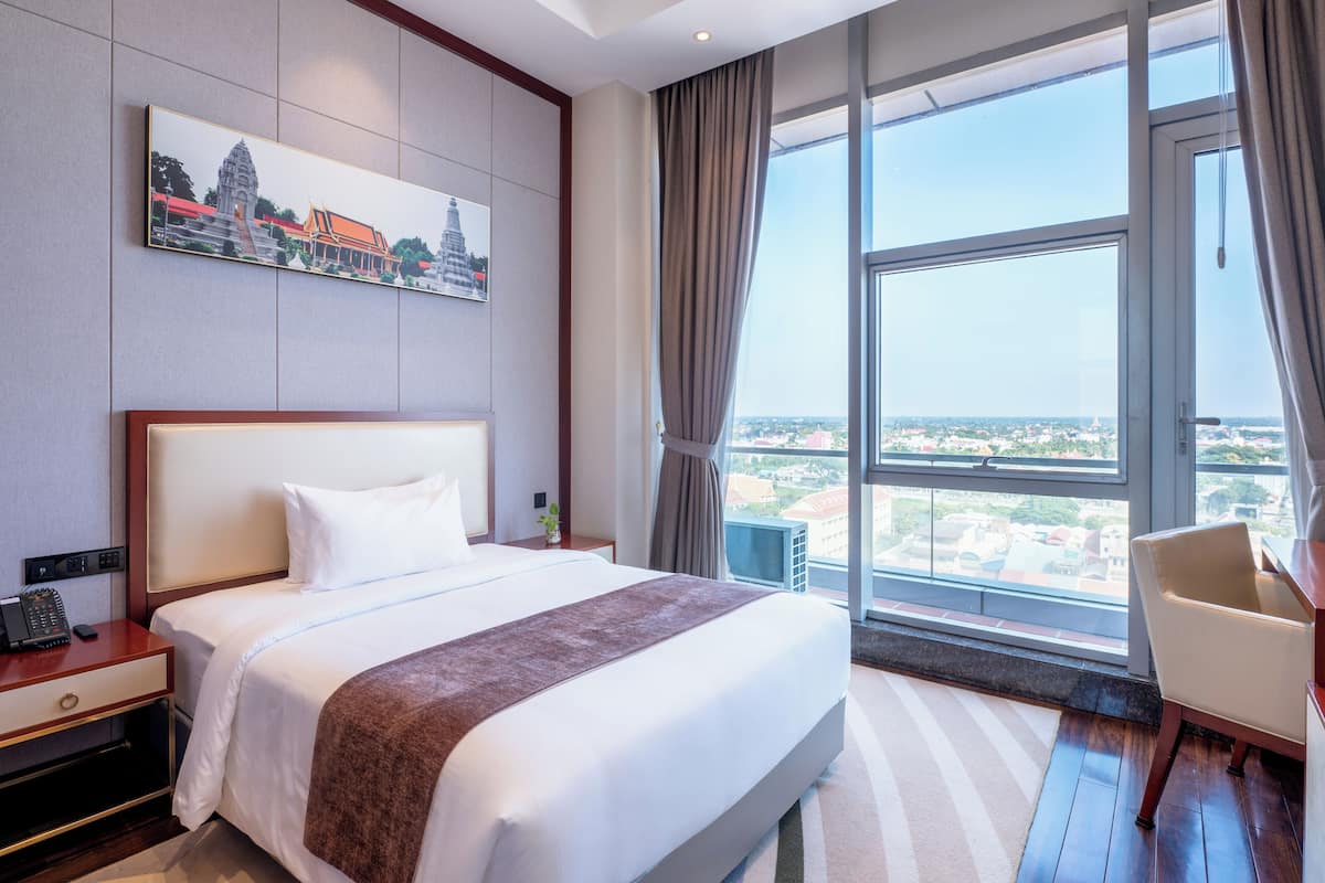 Een hotelkamer met een groot bed, een bureau, een stoel, een nachtkastje, een schilderij van een tempel en uitzicht op een stadslandschap door een groot raam.