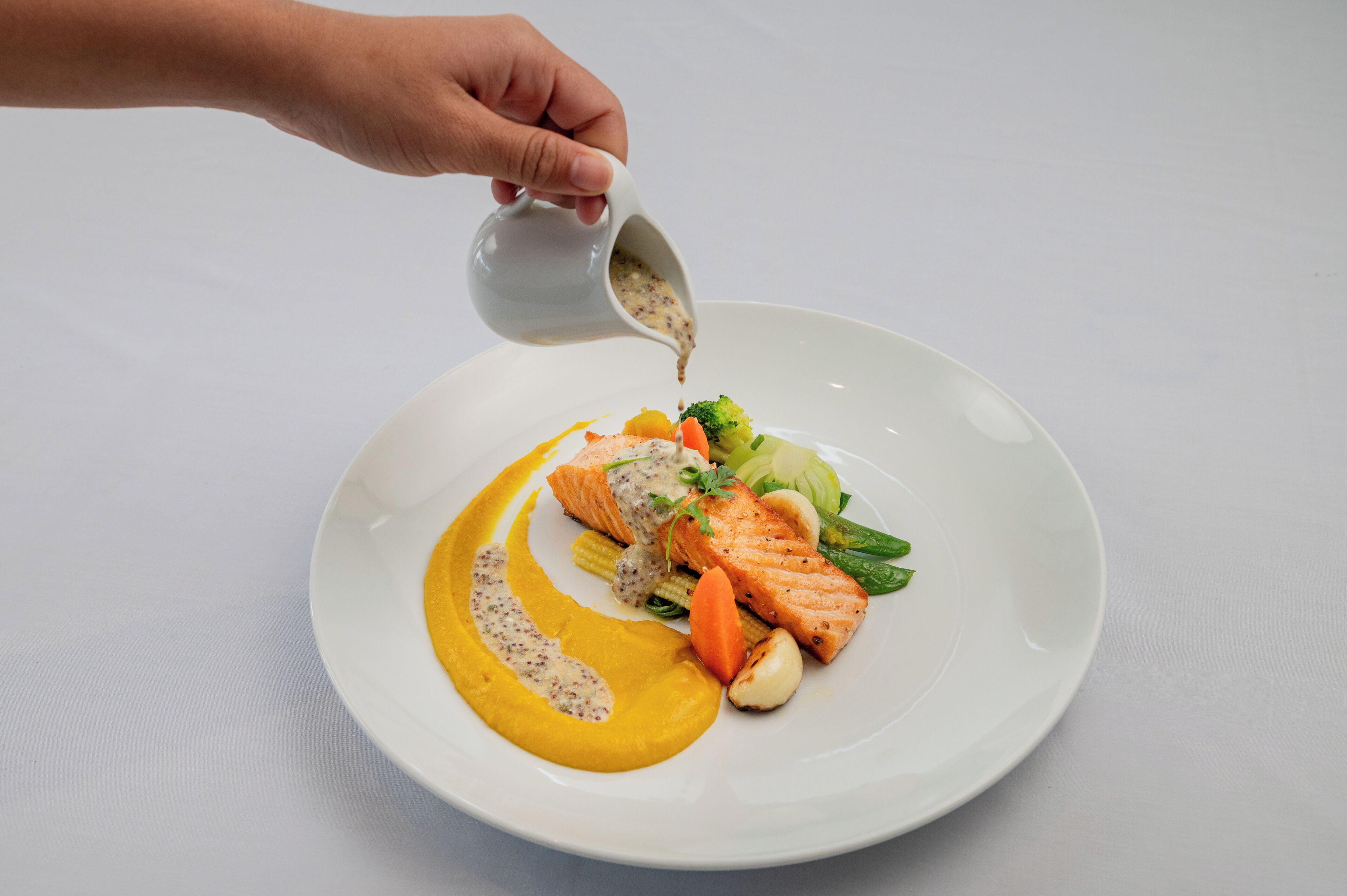 Una mà abocant salsa sobre un plat amb salmó, verdures i una salsa groga.