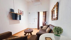 A living room with a sofa, a small round table, a TV mounted on the wall, a door, and a window with curtains. - Villa Paraíso Mango 4 (Jacó)