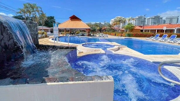 A swimming pool with a waterfall feature, lounge chairs, and a covered area with a red roof. - Villa Paraíso Mango 4 (Jacó)