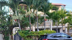 A black car parked in front of a gated entrance with a stone wall and steps leading up to a building with a red roof and multiple floors. - Cosy apartment unit in a triplex at less 100m from the beach with a common pool (Flic en flac)