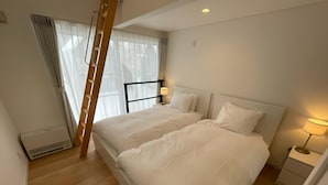 Un dormitorio con dos camas, una escalera, una ventana con cortinas, una mesita de noche con lámpara y un calentador fijado a la pared.