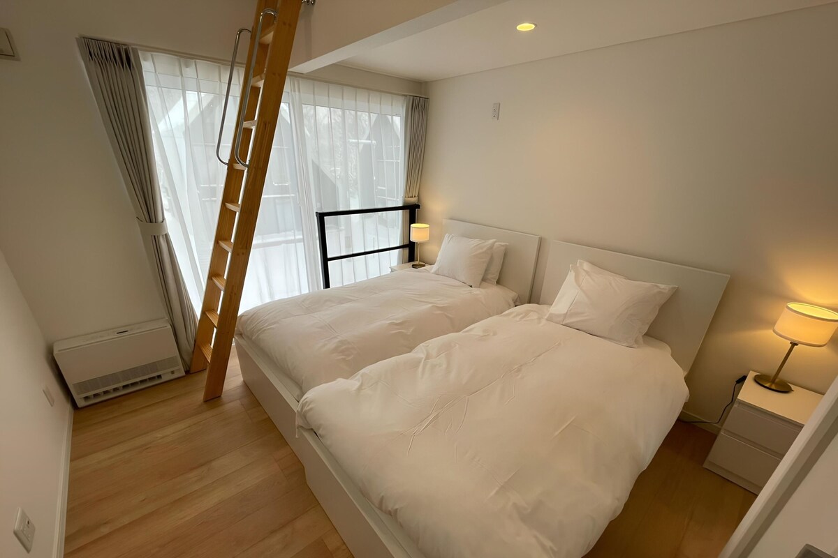 A bedroom with two beds, a ladder, a window with curtains, a bedside table with a lamp, and a wall-mounted heater.