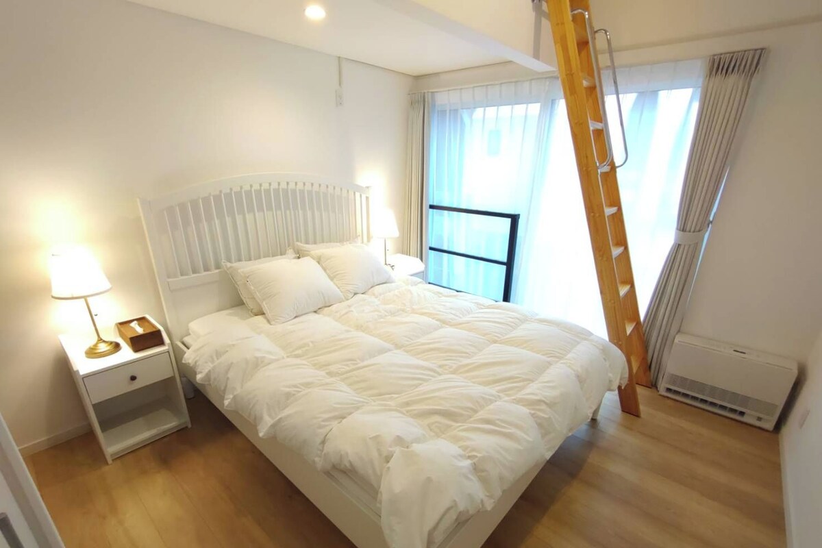 A bedroom with a bed, bedside table, lamp, window with curtains, and a ladder.