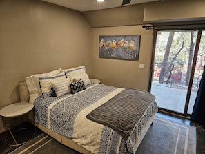Un dormitorio con una cama grande, una mesita redonda, un cuadro de pájaros en la pared y vistas a árboles por la ventana.