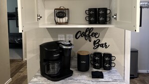 A kitchen area with a coffee station, including a Keurig coffee maker, a coffee grinder, and several black coffee mugs with the word "Coffee" on them.