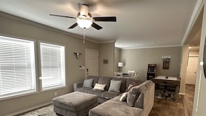 A spacious living room with a gray sectional sofa, two ottomans, a ceiling fan, and a dining area with a table and chairs.