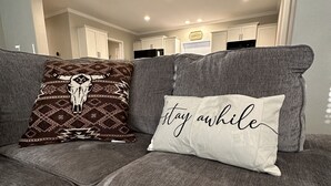 A grey sectional sofa with two decorative pillows, one with a cow design and the other with "stay awhile" text.