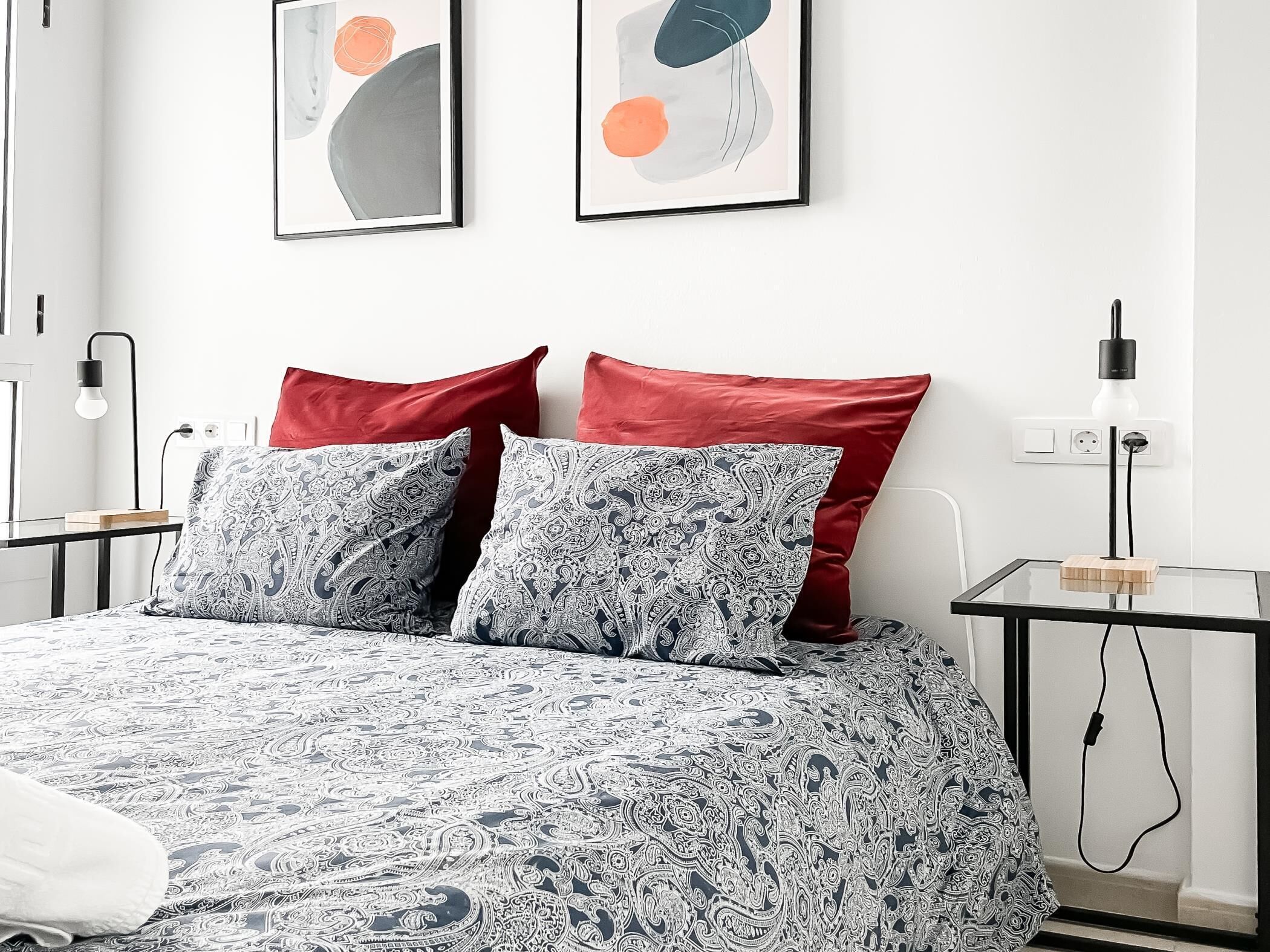A modern bedroom with a patterned bedspread, two red pillows, and abstract wall art.