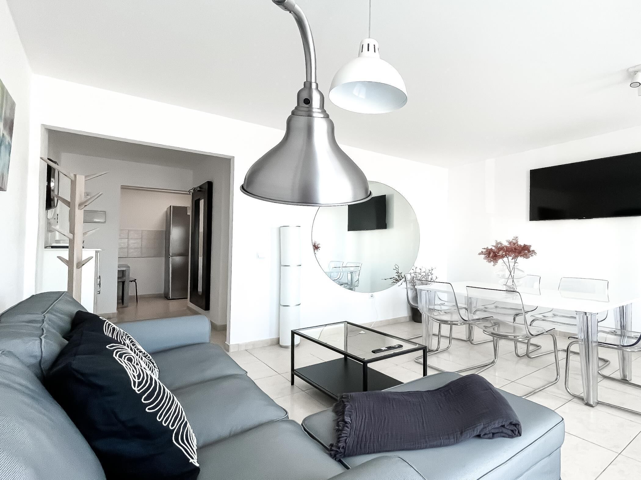 Un salon moderne avec un canapé gris, une table basse, un coin repas avec une table et des chaises en verre, et un grand miroir.