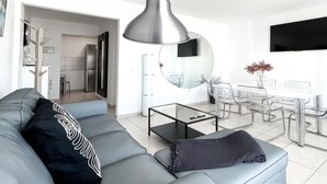 A modern living room with a grey sofa, a coffee table, a dining area with a glass table and chairs, and a large mirror. - Adela Homes by Best Holidays Fuerteventura (Corralejo)