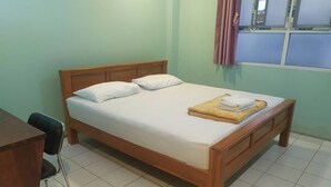 Ein ordentlich bezogenes Bett mit weißen Leinen und einem gefalteten Handtuch, ein hölzernes Kopfende, ein Schreibtisch mit Stuhl und ein Fenster mit Vorhängen.