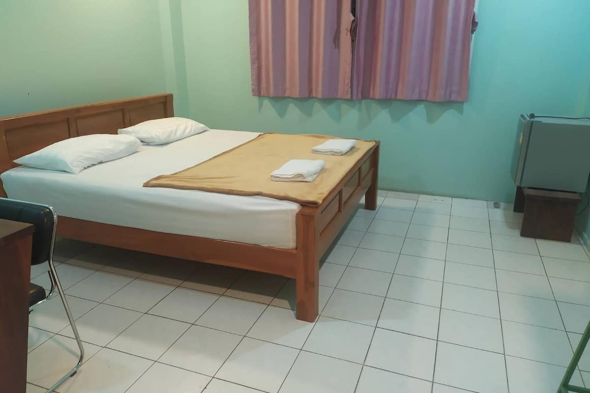 A single bed with a wooden frame, white bedding, and a beige blanket. There is a chair with a metal frame to the left of the bed.