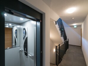 A modern elevator with a control panel and a glass door, a staircase with a metal railing, and a light fixture on the ceiling.