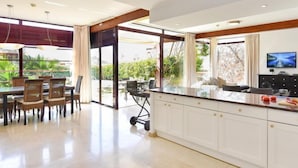A modern kitchen with white cabinets, a dining area with wicker chairs, and a large window overlooking greenery. - Villa Tauro 2 by Interhome (Mogan)