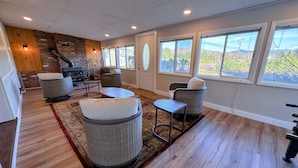A cozy living room with wicker furniture, a fireplace, and large windows offering a view of the outdoors. - Renovated home on snowmobile trail, firepit, hot tub, views,10 min from skiing, dogs welcome! (Twin Mountain)