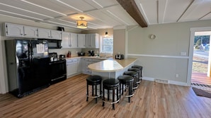 A modern kitchen with white cabinetry, a black refrigerator, a central island with stools, and a wooden floor. - Renovated home on snowmobile trail, firepit, hot tub, views,10 min from skiing, dogs welcome! (Twin Mountain)