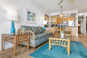 Ein Wohnzimmer mit einer Couch, Sesseln, einem Glastisch und einem Esstisch mit Stühlen. Im Hintergrund ist eine Küche mit Holzschränken und einer Mikrowelle zu sehen.
